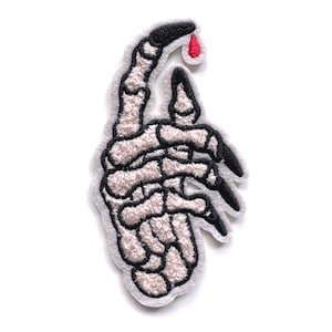 May include: A white skeleton hand with black outlines and long, sharp black fingernails. A single red drop of blood is on the tip of the index finger.