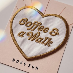 Puede incluir: Parche bordado en forma de corazón con el texto "coffee & a walk" en hilo dorado. El parche tiene un centro blanco y un borde dorado. El parche está sobre una tarjeta rosa con el nombre de la marca "NOVE SUN".