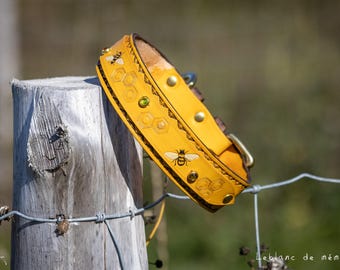 Puede incluir: Un collar de perro de cuero amarillo con diseños de abejas y panales. El collar tiene una hebilla y remaches dorados, y una gema verde. El collar está apoyado en un poste de madera desgastado.