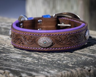 Puede incluir: Un collar de perro de cuero marrón con interior morado y conchos plateados decorativos. El collar presenta intrincados diseños tallados y está acentuado con costuras moradas. Una hebilla y una anilla en D plateadas completan el diseño.