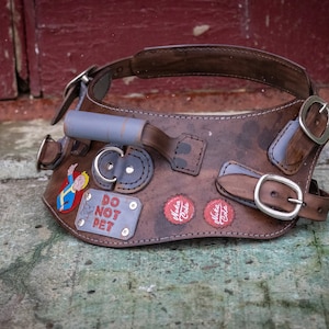 Pode incluir: Um arnês de cachorro de couro marrom com uma alça cinza, fivelas e patches. Os patches incluem um personagem de desenho animado, uma placa de "Não acariciar" e dois logotipos "Nuka Cola".