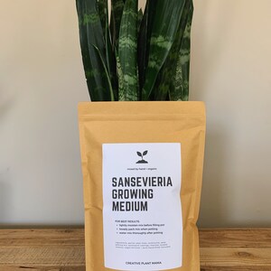 May include: A package of Sansevieria growing medium with a snake plant. The package is tan with a white label that reads "Sansevieria Growing Medium" and "Creative Plant Mama". The snake plant has long, green leaves with lighter green and yellow patterns.