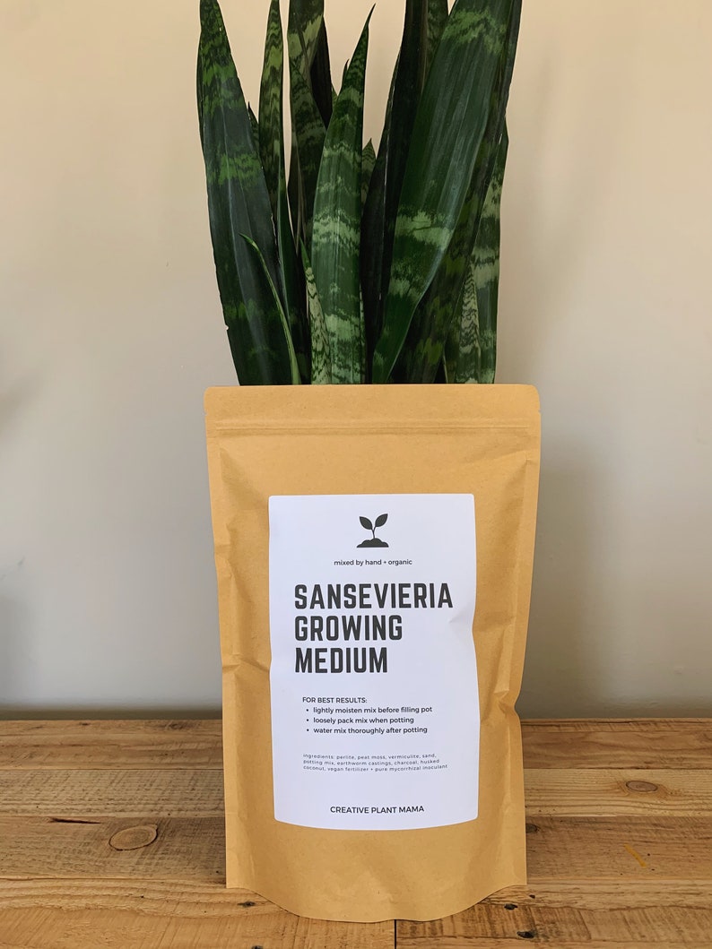 May include: A package of Sansevieria growing medium with a snake plant. The package is tan with a white label that reads "Sansevieria Growing Medium" and "Creative Plant Mama". The snake plant has long, green leaves with lighter green and yellow patterns.