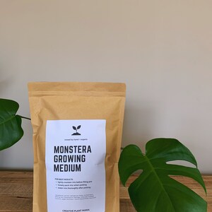 May include: A brown paper bag of "MONSTERA GROWING MEDIUM" with a white label. The label includes instructions and the text "CREATIVE PLANT MAMA". A large green Monstera leaf is on the right. The bag is on a wooden surface.