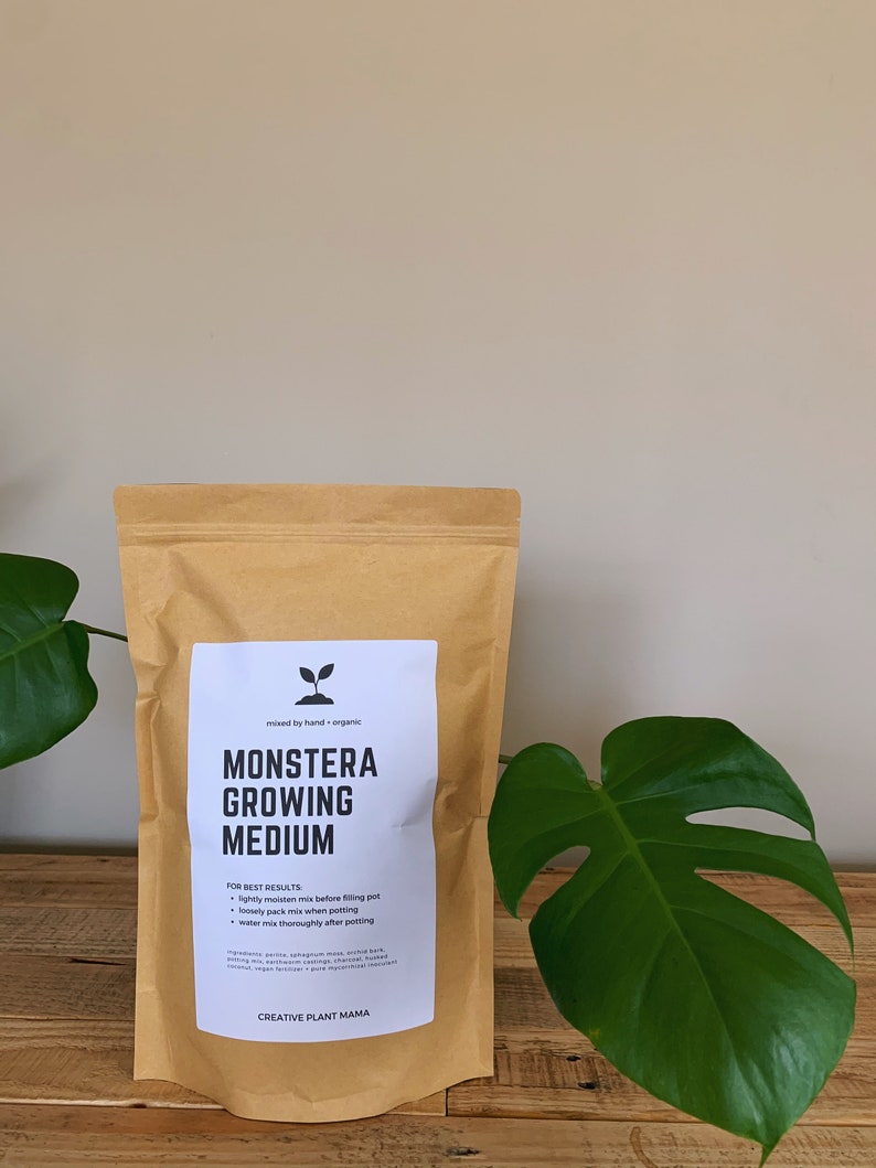 May include: A brown paper bag of "MONSTERA GROWING MEDIUM" with a white label. The label includes instructions and the text "CREATIVE PLANT MAMA". A large green Monstera leaf is on the right. The bag is on a wooden surface.