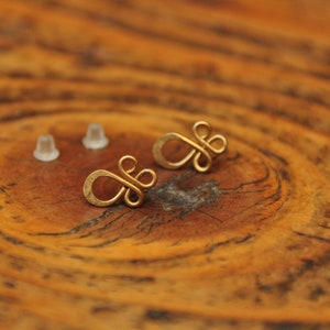 Puede incluir: Un par de pendientes de color dorado con un diseño simple anudado. Los pendientes están sobre una superficie de madera.