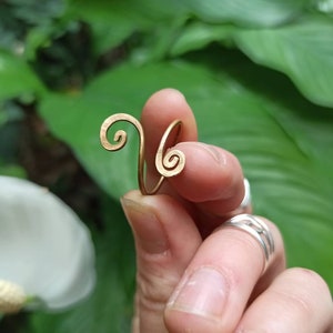 Puede incluir: Un anillo de alambre dorado con un diseño en espiral. El anillo está sostenido por una mano.