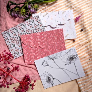 May include: A selection of patterned envelopes, including floral and dotted designs. One envelope displays a black and white poppy illustration. The envelopes are arranged on striped fabric, with sprigs of flowers.