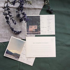 May include: Three postcards with scenic images and text. One postcard features a house with a blue door, another shows a landscape with a boat. The third postcard has lines for the recipient's address. Dried lavender sprigs are arranged on a linen cloth.