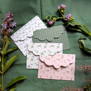May include: Four decorative envelopes with scalloped edges, each featuring a unique pattern. One is white with a floral design, another is green plaid, a third is light green with a floral pattern, and the last is pink with a botanical print. The envelopes are arranged on a green surface.