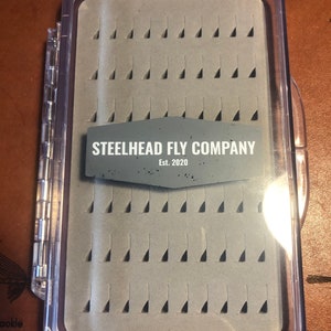 May include: A clear plastic fly fishing box with a grey foam insert and 64 compartments. The box has a label that reads "STEELHEAD FLY COMPANY Est. 2020".