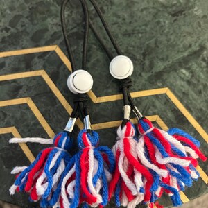 May include: Two red, white and blue tassel keychains with black elastic loops and white button closures.