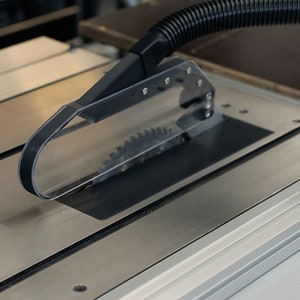 May include: A close-up of a table saw with a clear plastic blade guard and a black dust extraction hose attached. The saw is on a metal table with a silver finish.