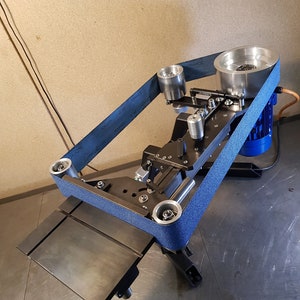 May include: A blue sanding belt runs around a black metal belt sander with a metal table and a blue electric motor. The belt sander has a metal table and a blue electric motor.
