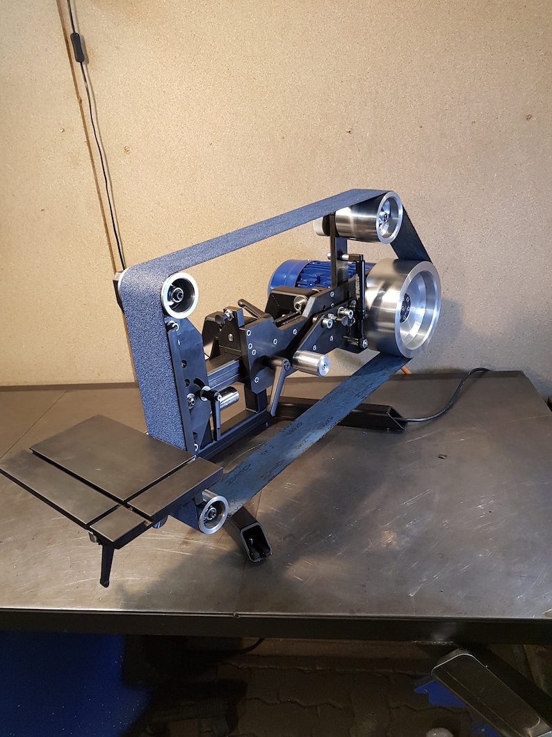 May include: A black metal belt sander with a blue sanding belt. The sander is mounted on a black metal stand with a metal work surface. The sander has a motor and a pulley system to move the belt.
