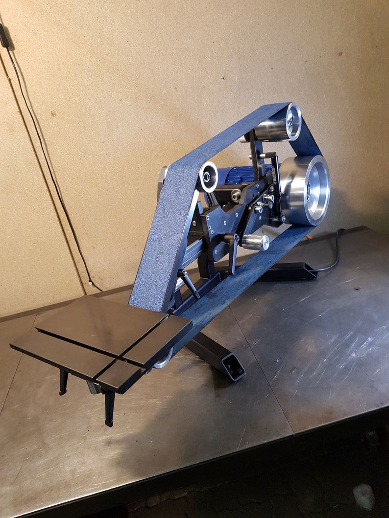 May include: A black metal belt sander with a blue motor and a black sanding belt. The sander is mounted on a black metal stand with adjustable legs. The sander is on a metal table.