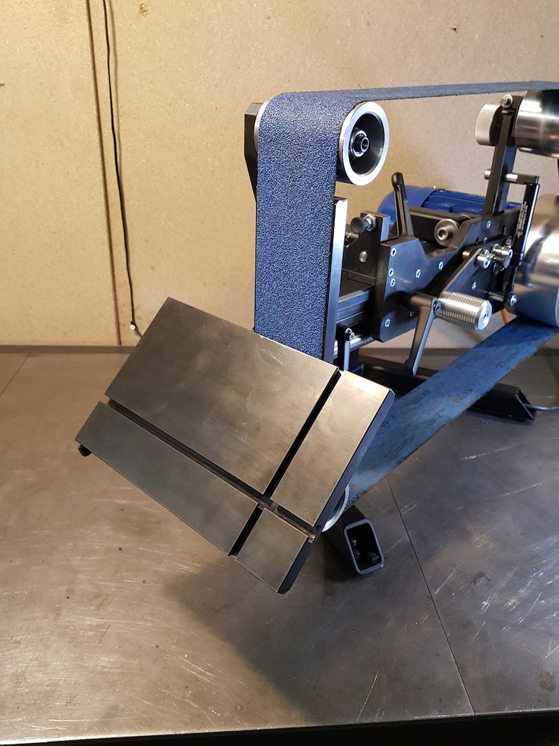 May include: A close-up of a black metal sanding table with a blue sanding belt running over it. The table is attached to a metal stand with a black motor and a blue switch.