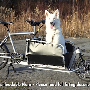 Könnte beinhalten: Ein silbernes Lastenrad mit einer schwarzen Transportbox, das einen weißen Hund transportiert. Das Fahrrad hat schwarze Reifen und einen Ständer. Der Hintergrund zeigt hohes, trockenes Gras. Der Text unten lautet: Downloadbare Pläne - Bitte lesen Sie die vollständige Angebotsbeschreibung.