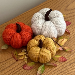 May include: Three knitted pumpkins in orange, cream, and mustard yellow, each featuring a dark brown stem. The pumpkins are round with ribbed textures and are arranged together with scattered small, colourful autumn leaves.
