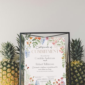 May include: A framed certificate of commitment with floral decorations. The certificate reads "Certificate of Commitment" and lists names and a date. Two pineapples flank the frame.