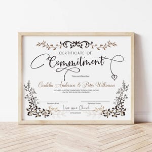 May include: A framed certificate with the words "Certificate of Commitment" in elegant script. The certificate includes names, a declaration, and space for signatures. Decorative floral elements in brown and black adorn the edges.