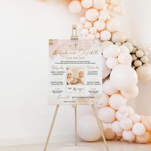 Könnte beinhalten: Eine hölzerne Staffelei zeigt ein Taufschild mit den Namen "Melinda und Elizabeth" in goldener Schrift. Das Schild enthält Fotos von zwei Babys, Details über ihre Taufe und ist mit einem rosa-goldenen Farbschema verziert. Ein Ballonbogen befindet sich im Hintergrund.