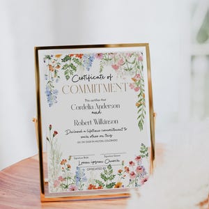 May include: A framed certificate with a gold border, featuring floral watercolour designs. The certificate reads "Certificate of Commitment" and includes names, a date, and location. The frame is displayed on a wooden surface.