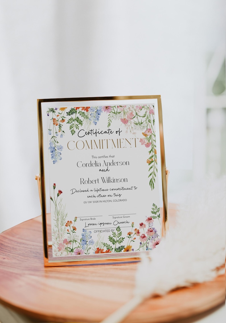 May include: A framed certificate with a gold border, featuring floral watercolour designs. The certificate reads "Certificate of Commitment" and includes names, a date, and location. The frame is displayed on a wooden surface.