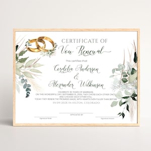 May include: A rectangular Certificate of Vow Renewal in a light wood frame. The white document features two gold rings, green eucalyptus leaves, and pampas grass illustrations. It includes printed text for names, dates, and location details.