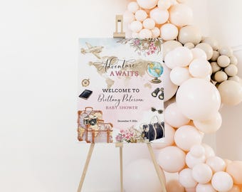May include: A baby shower welcome sign on a wooden easel. The sign features a world map, airplane, globe, and luggage illustrations with the text "Adventure Awaits" and "Welcome to Brittany Paterson Baby Shower".