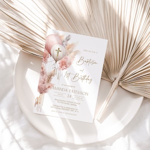 May include: A white invitation card with gold lettering announcing a baptism and 1st birthday party for Amanda Paterson. The card features a watercolor illustration of pink and white balloons with a gold cross, pampas grass, and other floral elements.