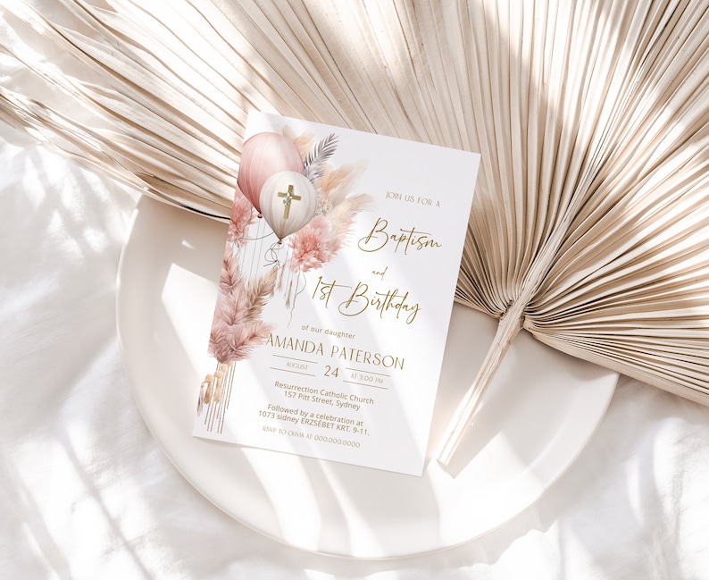 May include: A white invitation card with gold lettering announcing a baptism and 1st birthday party for Amanda Paterson. The card features a watercolor illustration of pink and white balloons with a gold cross, pampas grass, and other floral elements.