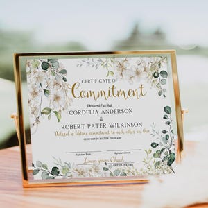 May include: A framed certificate with the words "Certificate of Commitment" in gold script. The certificate features floral and greenery designs in white and green. The names Cordelia Anderson and Robert Pater Wilkinson are listed on the certificate.