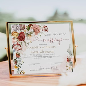 May include: A certificate of marriage printed on white paper, displayed in a gold metal frame. The document features floral illustrations in red, pink, and green, with text reading Certificate of Marriage, Cordelia Anderson and Pater Wilkinson.
