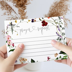 May include: A white prayer request card with floral accents and the words "prayer & request" in elegant script. The card features a cross and is lined for writing. The card is held by a person.