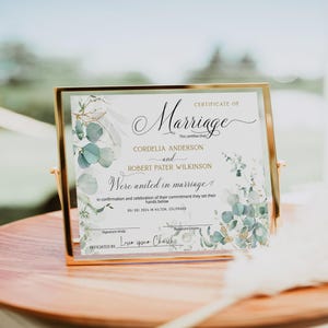 Könnte beinhalten: Eine gerahmte Heiratsurkunde mit dem Wort "Marriage" in eleganter Schrift. Die Urkunde enthält die Namen Cordelia Anderson und Robert Pater Wilkinson, mit floralen Akzenten in Grün und Gold. Das Datum ist der 05.09.2024.