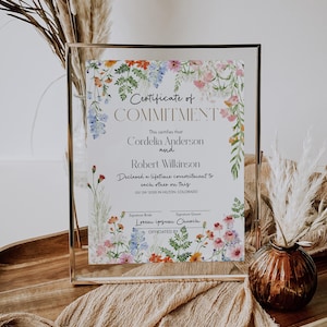 May include: A framed certificate, featuring the words "Certificate of Commitment" and floral decorations. The certificate is for Cordelia Anderson and Robert Wilkinson, dated 05/09/202X in Hilton, Colorado. The frame is gold.