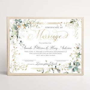 May include: A Certificate of Marriage document in a light wood frame, featuring elegant gold calligraphy and botanical illustrations of green eucalyptus leaves and gold accents. The text includes names, date, location, and signature lines.