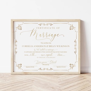 Pode incluir: Um certificado de casamento emoldurado com letras douradas e detalhes florais. O certificado diz "Certificate of Marriage" e lista os nomes Cordelia Anderson & Brian Wilkinson, com a data e o local.