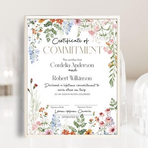 May include: A framed certificate with floral watercolour accents. The text reads "Certificate of COMMITMENT" above the names Cordelia Anderson and Robert Wilkinson. The certificate declares a lifetime commitment.