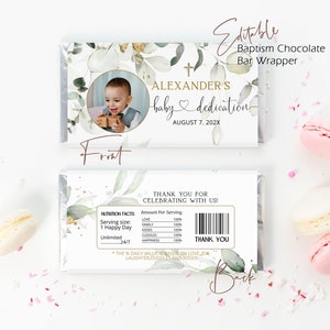 Puede incluir: Un envoltorio de barra de chocolate con diseño floral verde y blanco, con una foto de un bebé en una camisa blanca y una cruz dorada. El texto "ALEXANDER'S baby dedication AUGUST 7, 202X" está en la parte delantera del envoltorio. La parte posterior del envoltorio tiene un mensaje de agradecimiento y una etiqueta de información nutricional con el texto "1 Happy Day" y "Unlimited 24/7".