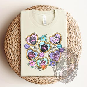 May include: A light green t-shirt with a design of paw prints in hearts. The hearts are surrounded by flowers and the words "Meow", "Show", and "Toy" are written on the hearts.