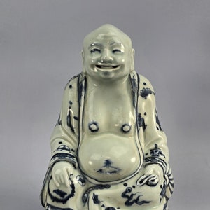May include: A white ceramic figurine of a smiling Buddha, wearing a blue and white robe, sitting with legs crossed. The figurine is decorated with blue floral patterns.