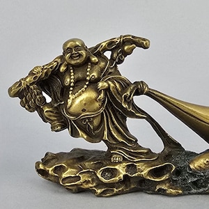 May include: A bronze-colored figurine of a smiling Buddha holding a large sack, standing on a textured base with a fish. The Buddha wears a necklace and flowing robes. The sculpture is detailed and has a polished finish.