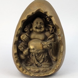 May include: A gold-colored metal sculpture of a smiling Buddha figure inside a hollowed-out egg shape.
