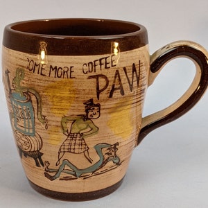May include: A brown ceramic coffee mug with a cartoon illustration of a person pouring coffee from a barrel. The mug has a brown rim and handle and the text "Come More Coffee Paw" is printed on the side.
