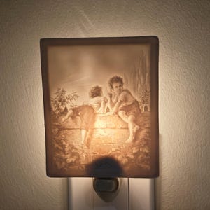 May include: A rectangular night light with a sepia-toned image of two children playing on a brick wall. The night light is plugged into a white wall outlet. The image depicts a classical scene.