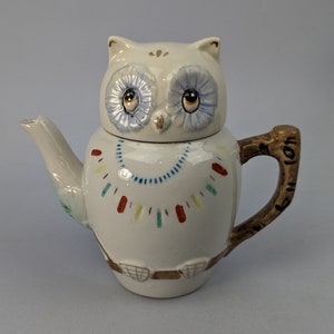 May include: White ceramic teapot shaped like an owl with blue eyes and a brown handle. The teapot has a colorful design around the body.