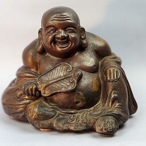 May include: A brown, smiling, Buddha statue sitting cross-legged with a fan in his hand.