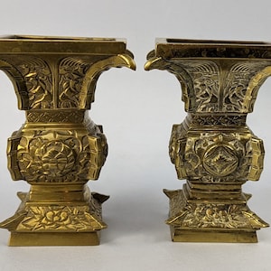 May include: A pair of gold-toned metal vases with intricate floral and geometric designs. The vases have a square base and a wide, flared opening.